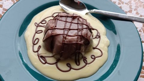 Ein brauner Schokoladenkloß liegt auf einer weißen Puddingcreme, dekoriert mit Schokolade auf einem blauen Teller.