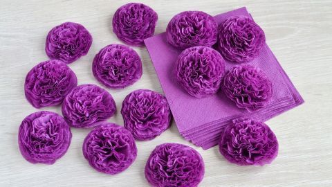 Bunte Papierblumen aus Servietten liegen auf einem hellen Tisch, umgeben von einer Gruppe gleichfarbiger Blüten.