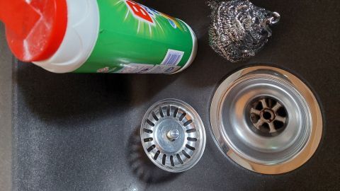 Ein Abflusssieb liegt neben Scheuerpulver und einer Stahlwolle in einem Spülbecken, bereit zum Reinigen.