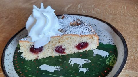 Ein Stück Kirschkuchen mit Dinkelmehl liegt auf einem dekorativen Teller, garniert mit Sahne und Puderzucker.