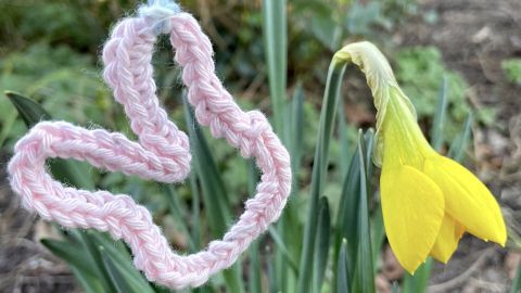 Ein gehäkelter Osterhase hängt an einem Faden neben einer gelben Blüte in einer grünen Gartenumgebung.