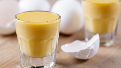 Zwei Gläser mit goldenem Eierlikör stehen auf einem Holztisch, umgeben von frischen Eiern und Eierschalen.