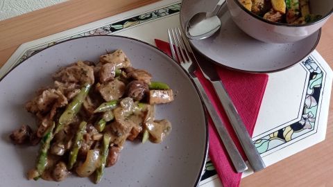 Schweinefilet-Ragout mit grünem Spargel liegt auf einem Teller, umgeben von einem bunten Tischsetting mit Besteck.