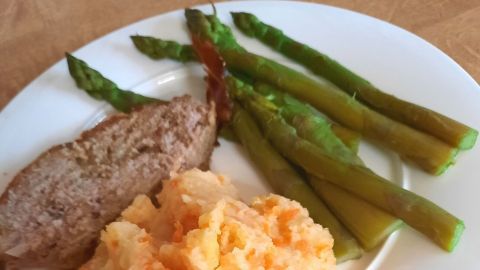 Saftiger Hackbraten liegt neben grünem Spargel und cremigem Kartoffel-Karottenstampf auf einem weißen Teller.