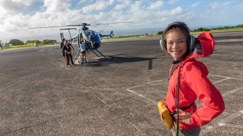 Eine junge Frau mit Kopfhörern lächelt in einem roten Anorak und steht vor einem Helikopter auf dem Flughafen.