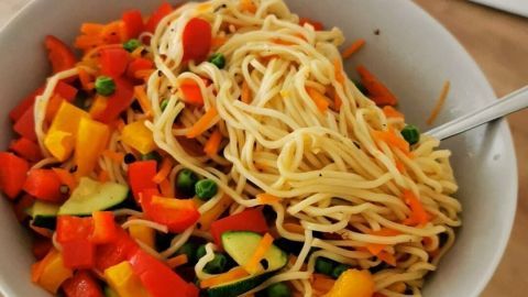 Eine Hand hält eine Schüssel mit vegetarischen Mienudeln und buntem Gemüse in einer hellen, modernen Küche.