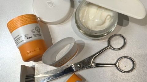 Ein offener Cremetopf steht neben leeren Tuben und einer Schere, während Reste von Kosmetikprodukten auf einem Tisch liegen.