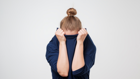Eine Person mit einem blauen Shirt zieht es über ihr Gesicht, als ob sie sich verstecken möchte, vor einem neutralen Hintergrund.