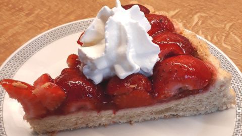 Eine Tortenscheibe mit Erdbeeren und Sahne steht auf einem weißen Teller, umgeben von einer Holzoberfläche.