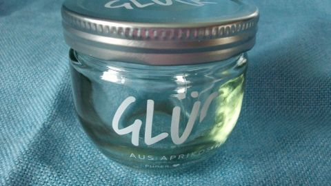 Ein Glas mit der Aufschrift "GLÜK" steht auf einem blauen Stoff, während es mit transparentem Inhalt gefüllt ist.