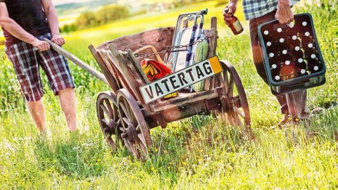 Ein Bollerwagen wird von zwei Männern im grünen Feld geschoben, beladen mit Sachen für den Vatertag, einer hält ein Bier.