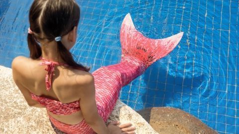 Ein Mädchen mit einer rosa Meerjungfrauenflosse sitzt am Poolrand und betrachtet das Wasser.