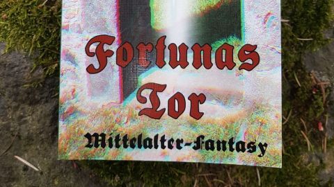 Ein Buch mit dem Titel „Fortunas Tor“ liegt auf einem moosbedeckten Stein in einer natürlichen Umgebung.