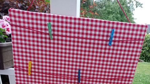 Eine rot-weiß karierte Pinnwand hängt draußen; bunte Klammern halten Notizen und Bilder im Freien fest.
