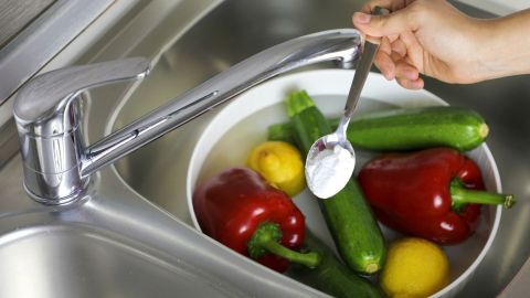Eine Hand hält einen Löffel mit weißem Pulver über eine Schüssel mit Gemüse im Wasser, um Obstfliegen zu verhindern.