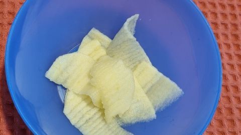 In einer blauen Schüssel liegen sicher geschnittene Obststücke, bereit für das Essen von Kleinkindern, auf einem orangefarbenen Untergrund.