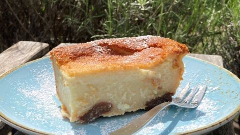 Ein Stück Far Breton liegt auf einem blauen Teller, umgeben von frischem Grün, leicht mit Puderzucker bestäubt.