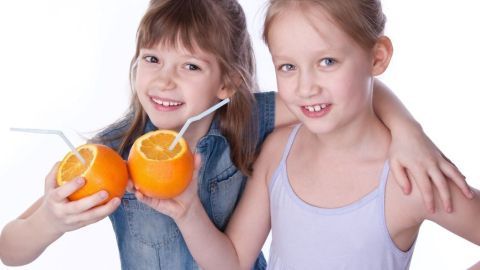 Zwei Mädchen halten orangefarbene Orangen mit Strohhalmen und lächeln fröhlich in einem hellen, neutralen Hintergrund.
