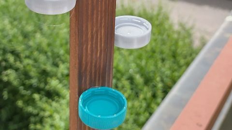 Ein Holzpfahl trägt bunte Flaschenverschlüsse, die als Tränke für Insekten dienen, in einer grünen Gartenumgebung.