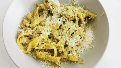 Bandnudeln liegen in einer schlichten Schüssel, mit Sahne-Pesto und geriebenem Käse garniert. Landschaftsfond aus Holz.