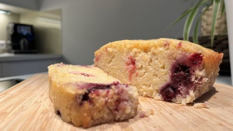 Ein Grießkuchen mit Beeren liegt auf einem Holzbrett, während ein Stück davon bereits abgeschnitten ist.