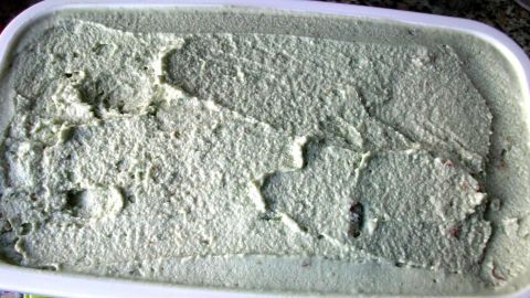 Eine cremige, grüne Avocado-Walnuss-Eiscreme liegt in einem weißen Behälter auf einem Tisch, umgeben von einer bunten Marmoroberfläche.
