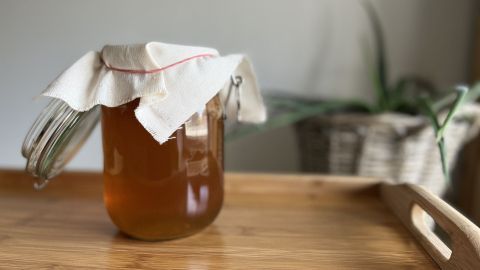 Ein Glas mit fermentiertem Tee-Getränk, abgedeckt mit einem Tuch, steht auf einem Tisch in einer hellen Umgebung.