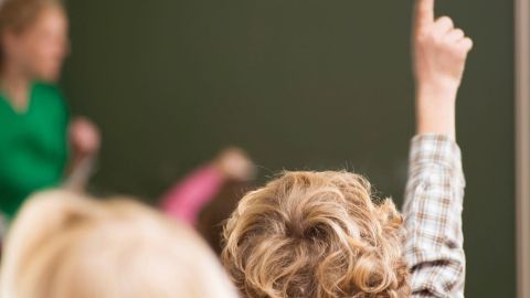 Ein Kind mit Locken hebt die Hand im Klassenraum, während eine Lehrerin im Hintergrund den Unterricht leitet.