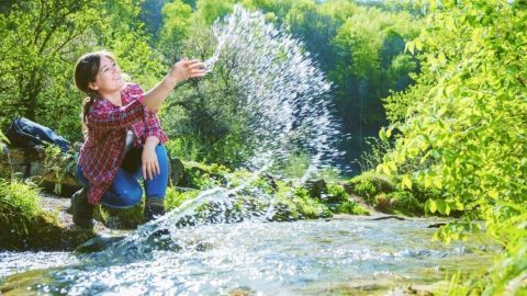Ein Mädchen spielt im Wasser eines klaren Flusses und spritzt fröhlich Wasser in die grüne, natürliche Umgebung.