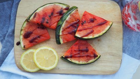 Auf einem Holzbrett liegen gegrillte Wassermelonenstücke, garniert mit einem süßen Sirup, neben zwei Zitronenscheiben.