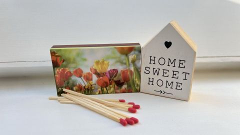 Ein Haus im Miniaturformat steht neben einer Streichholzschachtel, während bunte Blumen auf der Vorderseite zu sehen sind.
