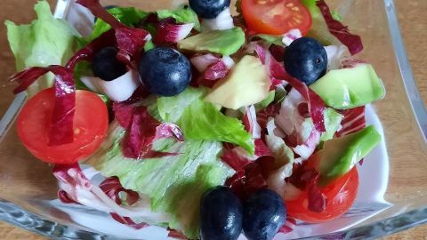 Frischer Salat mit Beeren, Tomaten und Avocado wird in einer klaren Schüssel auf einem Holzuntergrund serviert.