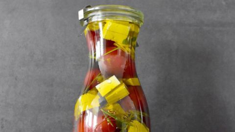 Ein Glas mit eingelegten Cherrytomaten, Feta und Kräutern steht auf einer dunklen Oberfläche neben frischem Knoblauch.