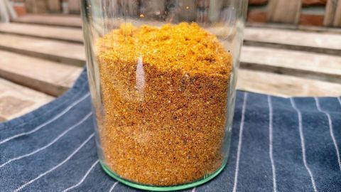 Eine Gewürzmischung für Nudeln befindet sich in einem Glas auf einem karierten Tuch, umgeben von Holzflächen.