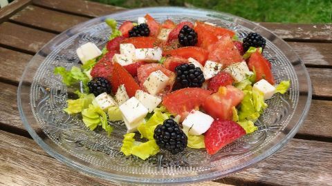 Frischer Salat mit Tomaten, Erdbeeren und schwarzen Beeren in einer gläsernen Schüssel, serviert auf einem Holztisch.