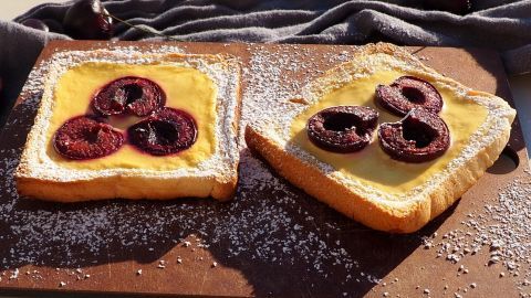 Zwei Joghurt-Toasts liegen auf einem Holzbrett, belegt mit Früchten und Puderzucker, in einer sonnigen Umgebung.