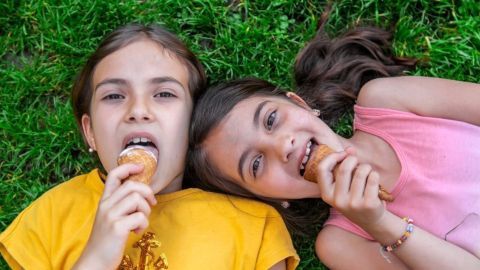 Zwei Mädchen liegen auf dem Gras und essen Eis, während sie lächeln und Spaß haben.
