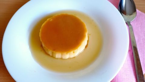 Eine runde Crème Caramel auf einem weißen Teller liegt in einer goldenen Karamellsoße, umgeben von einem Holzuntergrund.