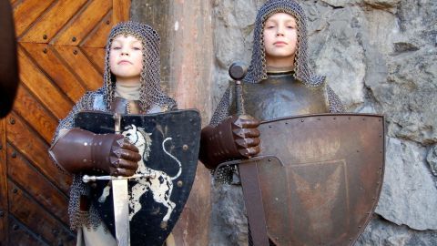 Zwei Ritter-Kinder stehen stolz vor einer steinernen Wand, jeweils mit Schwert und Schild, in voller Rüstung.