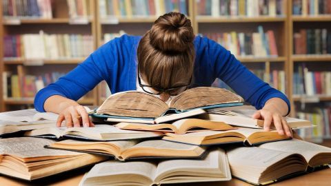 Eine Person mit Brille und blauer Kleidung beugt sich erschöpft über zahlreiche Bücher auf einem Tisch in einer Bibliothek.