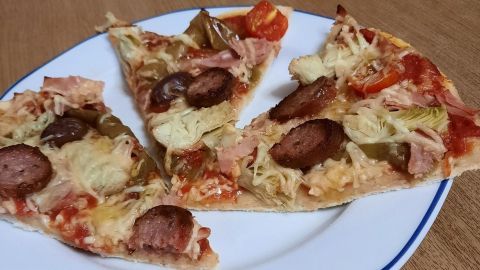 Drei Stücke Pizza mit Artischocken und Kümmelwurst liegen auf einem weißen Teller, umgeben von einem holzfarbenen Tisch.