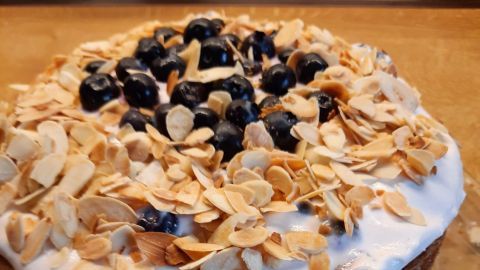 Eine Heidelbeertorte mit Mandeln steht auf einem grazilen Teller. Sie hat eine cremige Schicht und ist mit gerösteten Mandeln und frischen Heidelbeeren dekoriert.