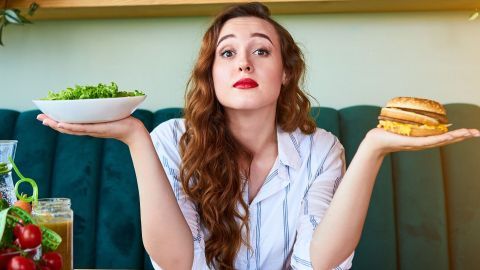 Eine Frau hält in jeder Hand eine Schüssel mit Salat und einen Burger, skeptisch und nachdenklich blickend.