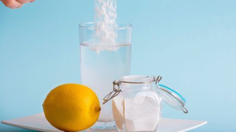 Ein Löffel Natron wird in ein Glas Wasser gegeben, während eine Zitrone und ein offenes Glas Zucker auf einem Teller stehen.