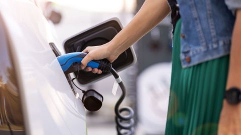Eine Person steckt ein Ladekabel in ein Elektroauto, um es aufzuladen, während im Hintergrund eine Ladesäule sichtbar ist.