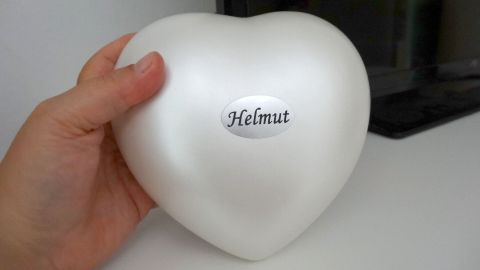 Eine Hand hält eine herzförmige Urne mit dem Namen "Helmut" darauf, im Hintergrund ein Monitor.