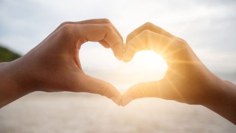 Hände formen ein Herzsymbol, während ein Sonnenstrahl im Hintergrund aufgeht, was das Thema Selbstliebe verstärkt.