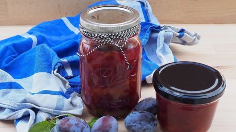 In einem Glas und einem kleinen Behälter ist ein köstlicher Zwetschgenröster ohne Alkohol neben frischen Zwetschgen und einem blauen Tuch.
