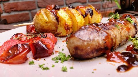 Gegrillte Kartoffelspieße und Bratwurst liegen saftig auf einem Teller, umgeben von frischen Tomaten und Kräutern.