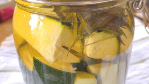 Ein Glas mit eingelegten Zucchini und Kräutern steht auf einem weißen Tuch und reflektiert das Licht aus der Umgebung.